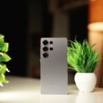 Modern smartphone standing on a desk with green potted plants in a warm indoor setting.