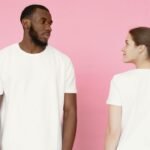 A man and woman in white t-shirts stand against a pink backdrop.