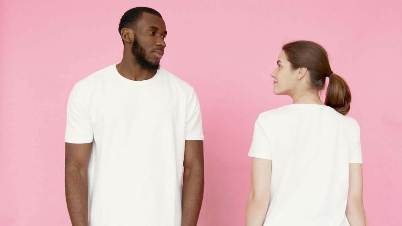 A man and woman in white t-shirts stand against a pink backdrop.